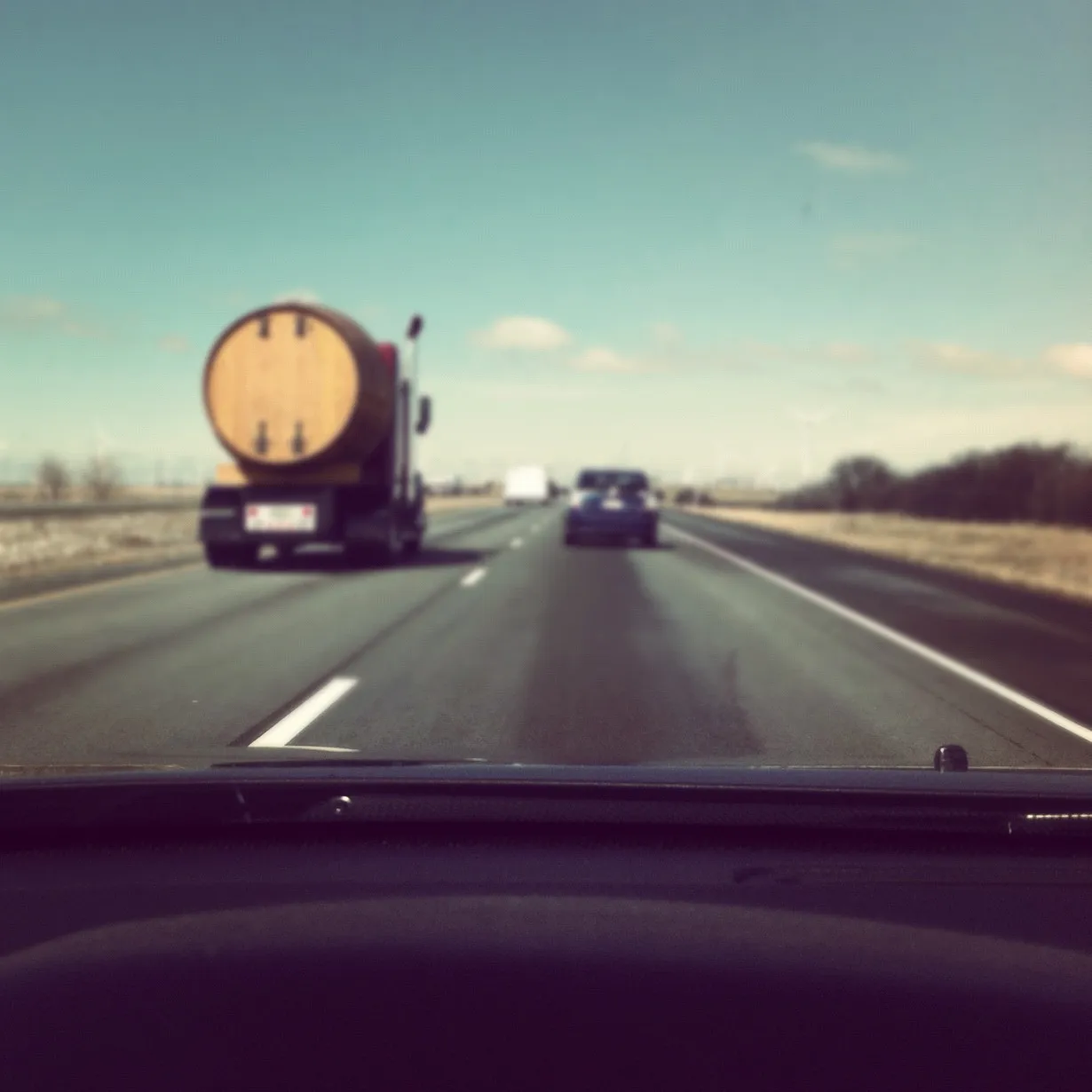 View from a car driving on a highway behind a truck carrying a large barrel-shaped container, under a clear blue sky.