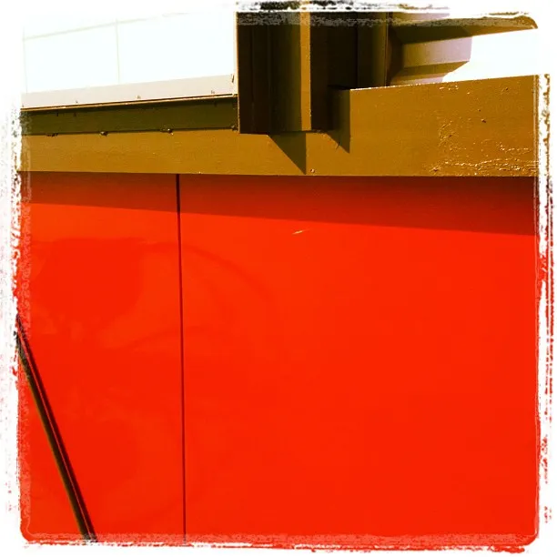 Bold red and olive green architectural detail featuring a metal panel with vertical seams and an overhanging structural element.