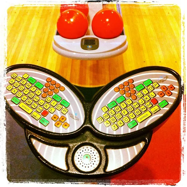 Colorful dual-keypad bowling score console with rounded keys, positioned in front of two red bowling balls on a ball return rack.
