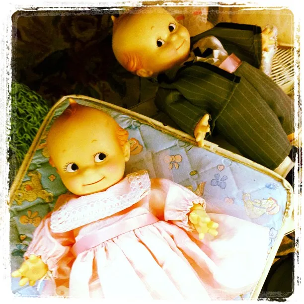 Two vintage Kewpie dolls with wide eyes and smiling faces; one dressed in a pink dress and the other in a striped suit, posed on a patterned cushion.
