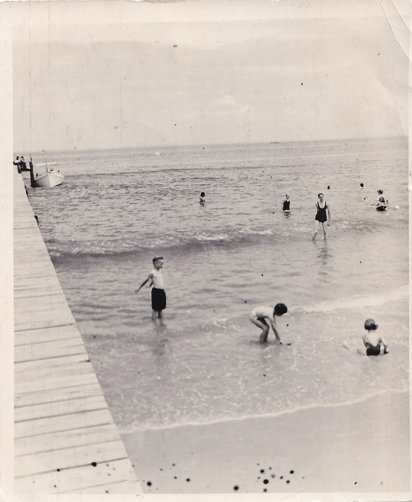 Vintage black and white photograph of children swimming and wading in shallow water near a wooden dock, with a small boat moored in the calm background