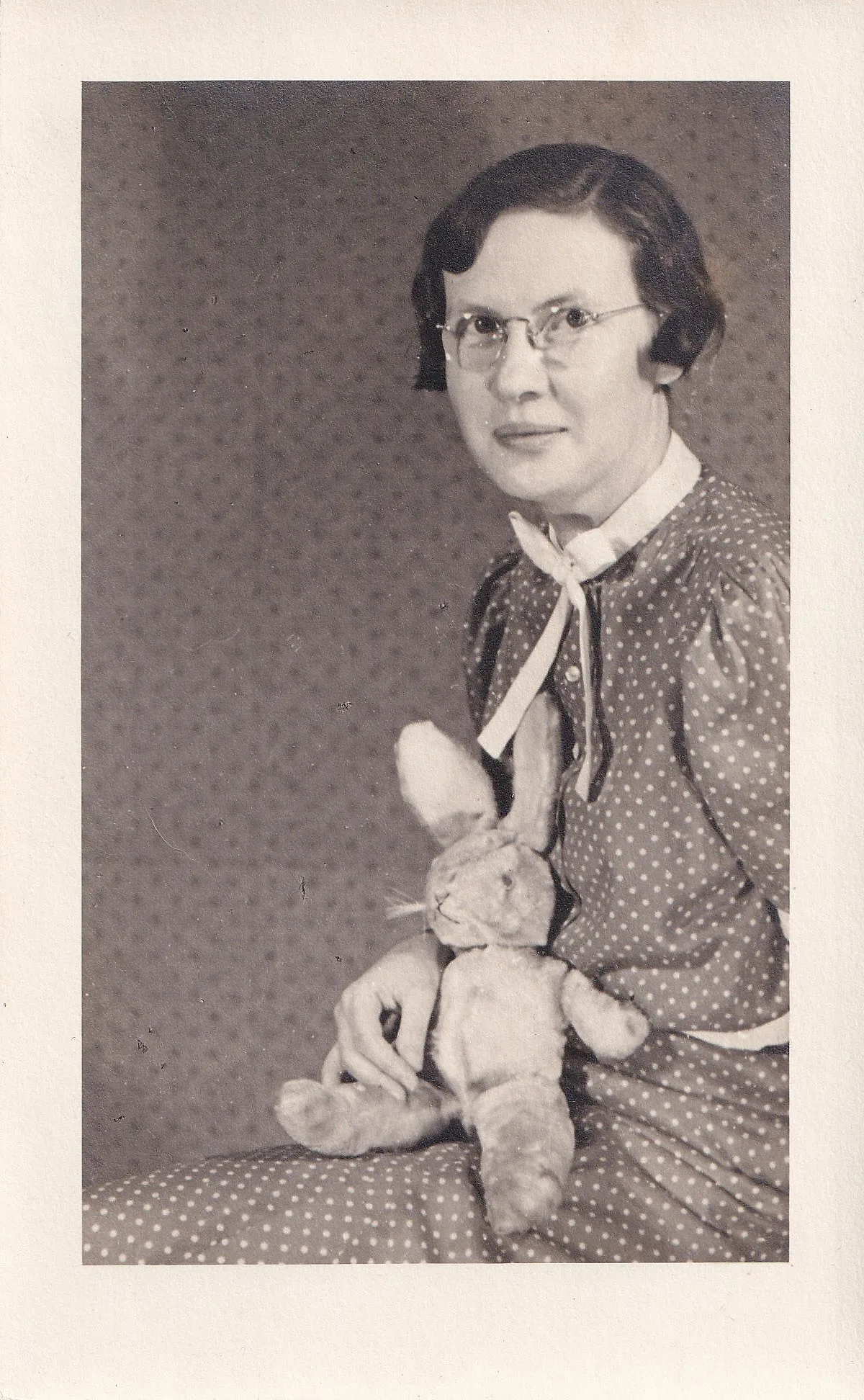 Formal black and white studio portrait of a middle-aged woman in wire-rimmed glasses and a polka-dot dress, holding a stuffed rabbit toy in her lap