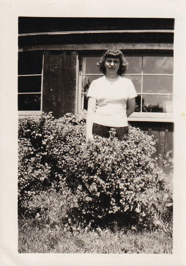 Vintage black and white snapshot of a woman in a white t-shirt standing behind a large flowering bush in front of a wooden building
