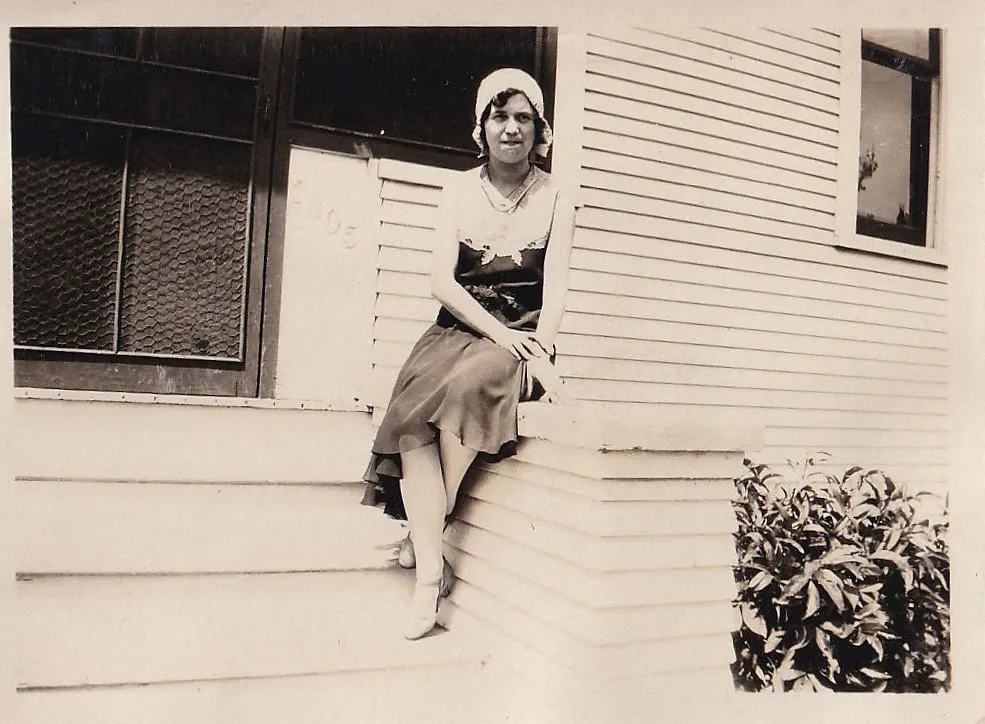 Vintage black and white photograph of a young woman in a cloche hat sitting on the front porch steps of a clapboard house, house number 305 visible on the post beside her