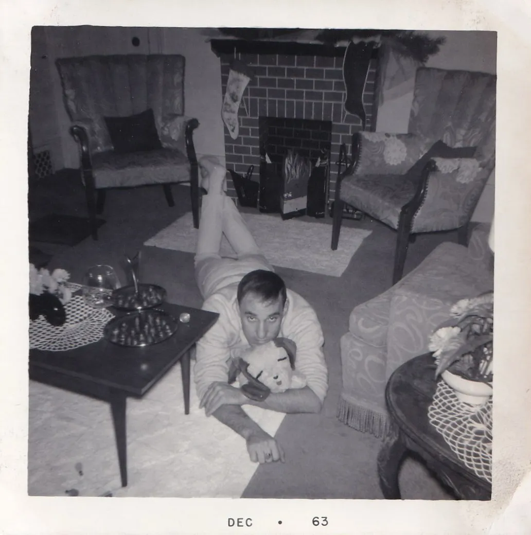 Black and white snapshot of a man lying on a living room floor holding a stuffed dog toy, Christmas stockings hung on the brick fireplace behind him, dated December 1963