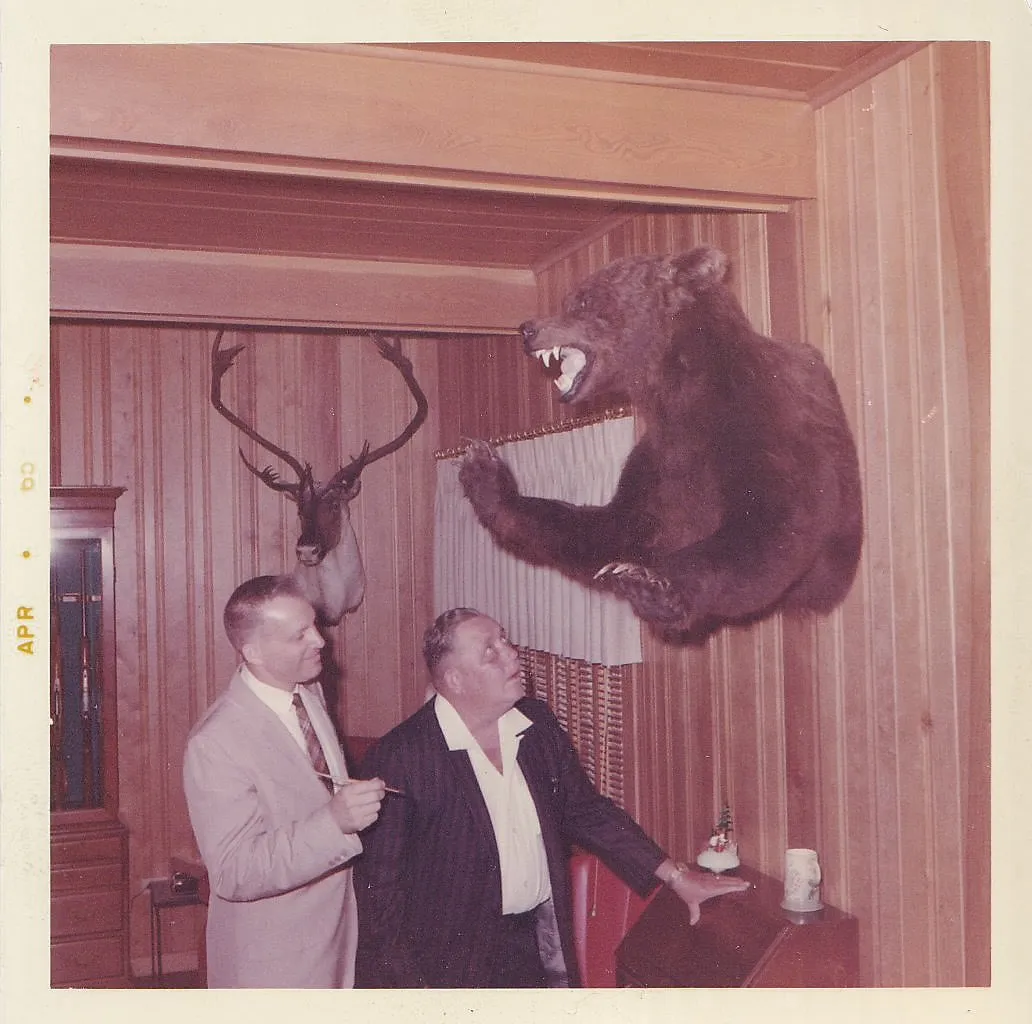 Color snapshot of two men in a wood-paneled room standing beneath a mounted bear taxidermy with open mouth and raised paws, deer antlers on the wall behind, April 1960