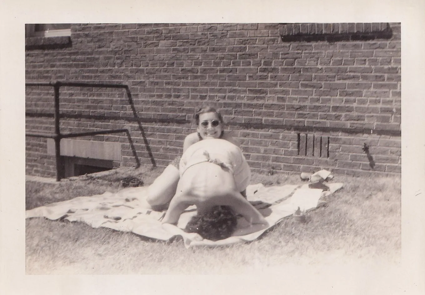 Vintage black and white photograph of a woman in sunglasses and a man sunbathing side by side on a blanket against a brick wall, the woman looking up at the camera