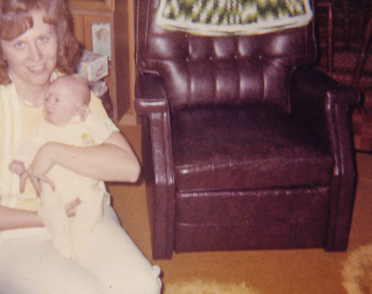 Found color snapshot of a young woman with red hair holding a newborn baby while seated on the floor beside a brown leather recliner in a 1970s domestic interior