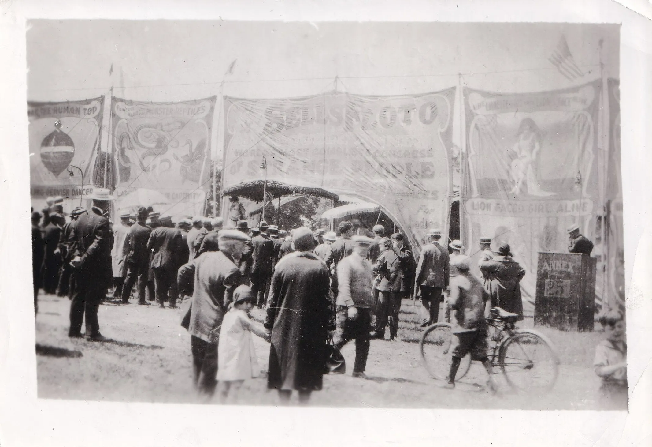 Vintage black and white photograph of a crowd gathered at a carnival sideshow, painted banner signs reading 'Lion Faced Girl Alive' and 'The Human Top' visible above them, early twentieth century