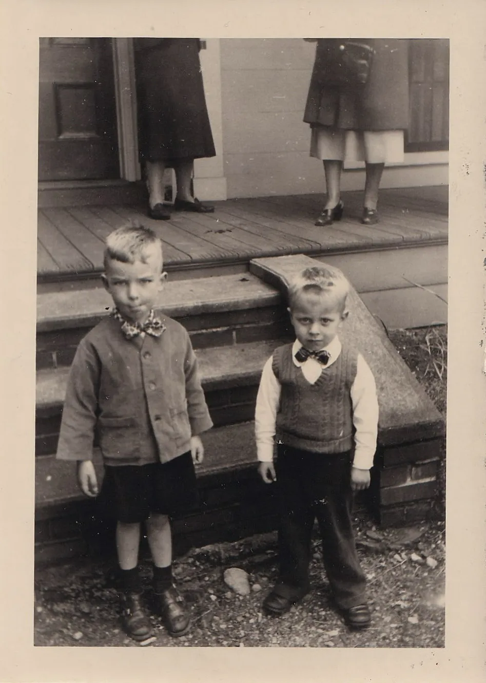 Vintage black and white photograph of two young boys in formal bow ties standing before a house porch, two adult women visible only from the waist down in the background