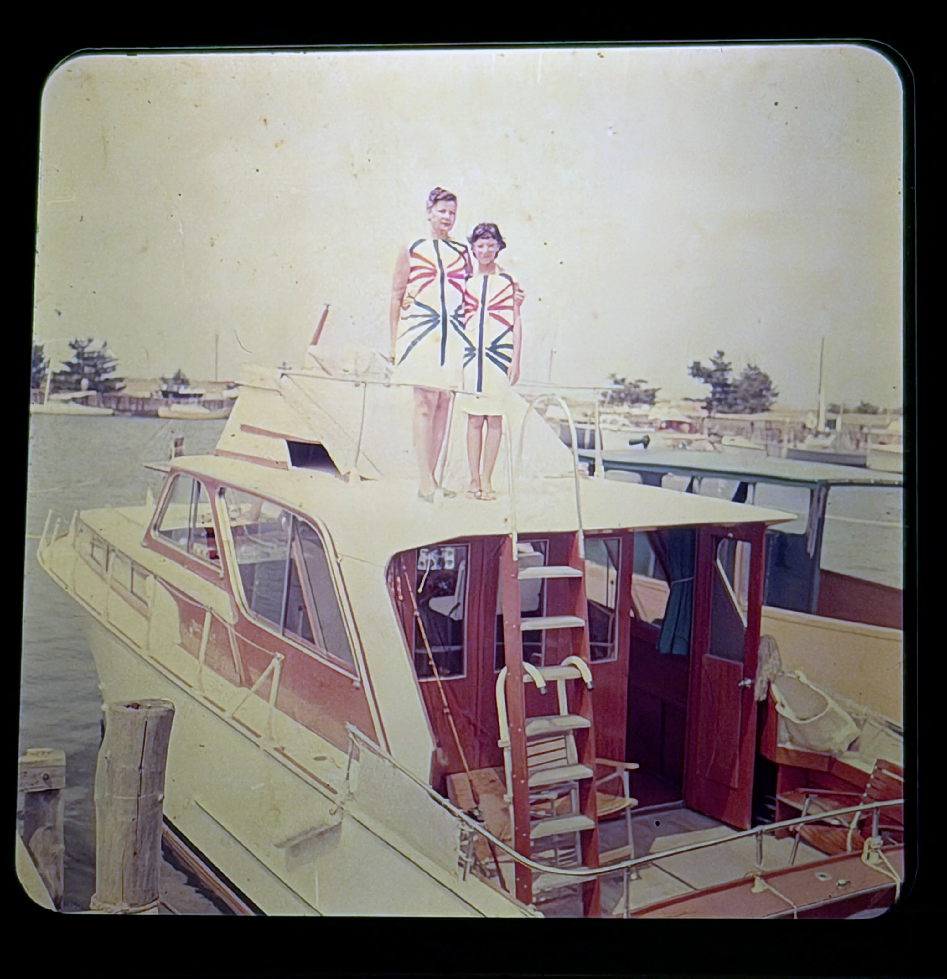 Vintage color photograph of two women in matching patterned dresses standing on cabin cruiser flybridge at marina"