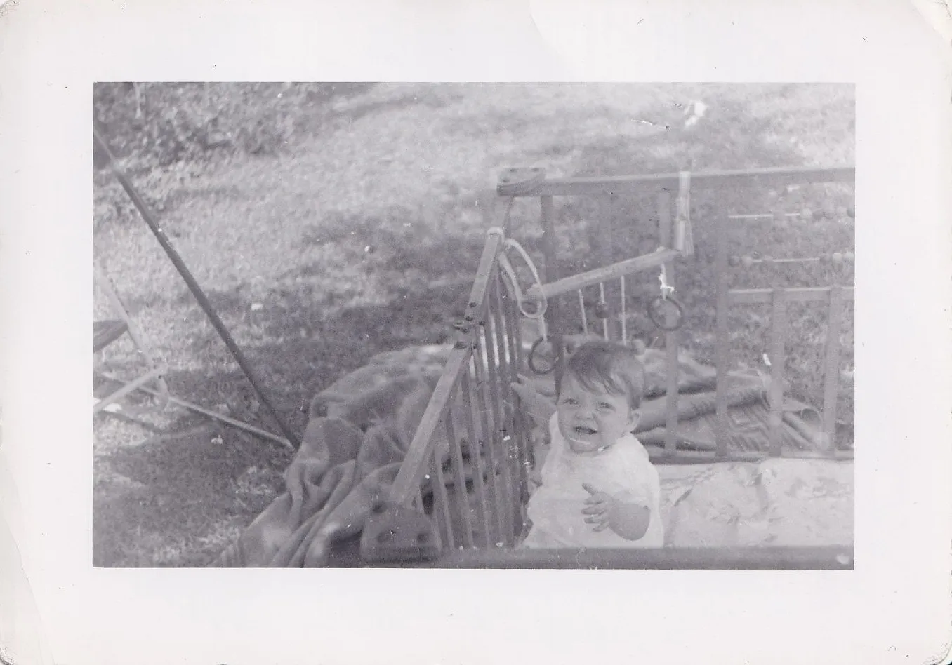 Vintage black and white photograph of a smiling baby seated outdoors in a wooden playpen, backyard grass and garden equipment visible in the background