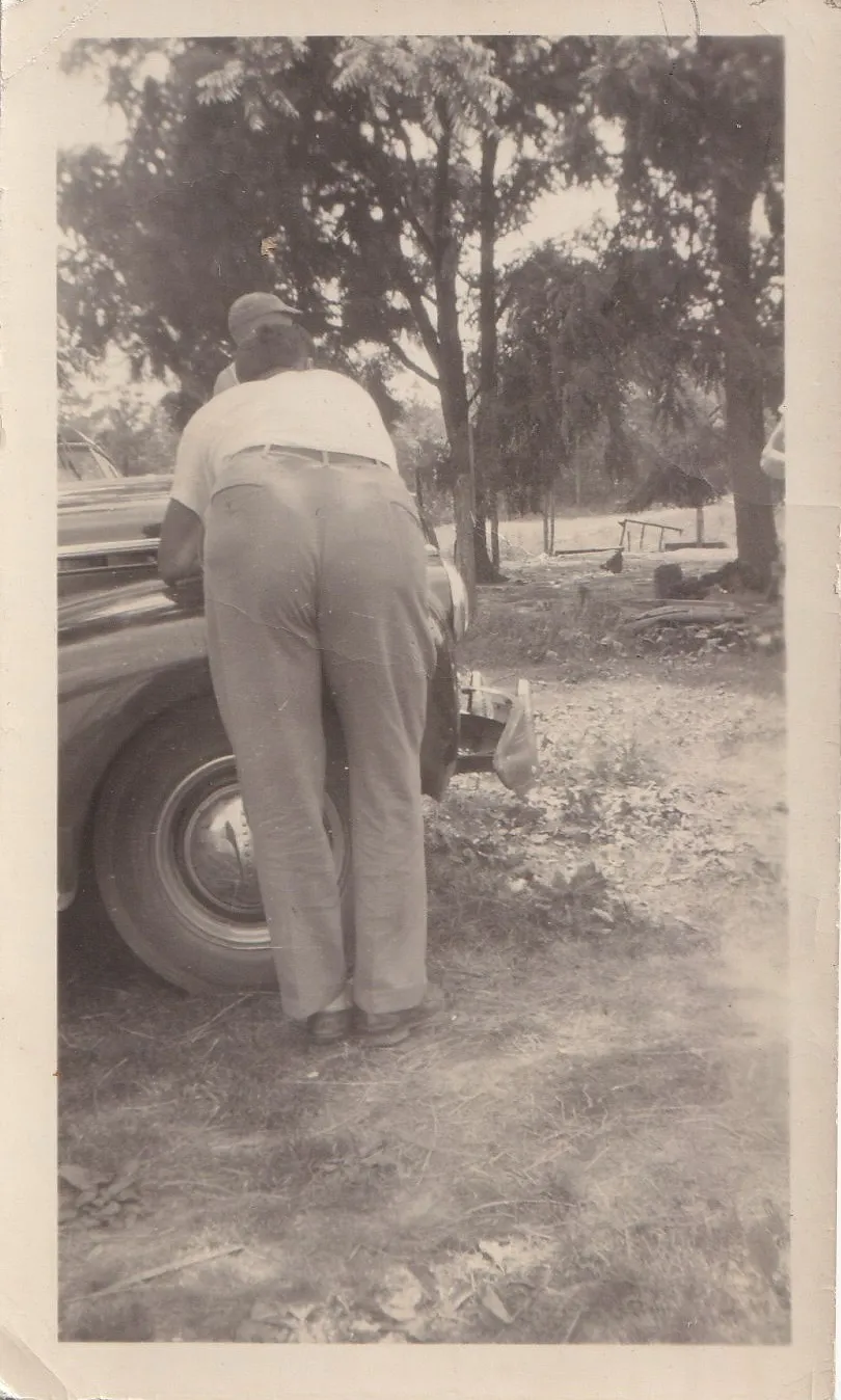 Vintage black and white photograph of a man photographed from behind, standing at the rear of a large vintage American car on a dirt lot surrounded by trees