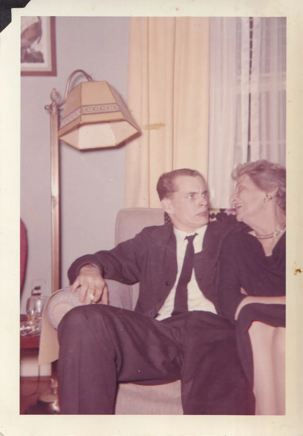 Found color snapshot of a young man in a dark suit and an older woman in pearls seated side by side on a sofa, he staring away stiffly while she leans toward him laughing