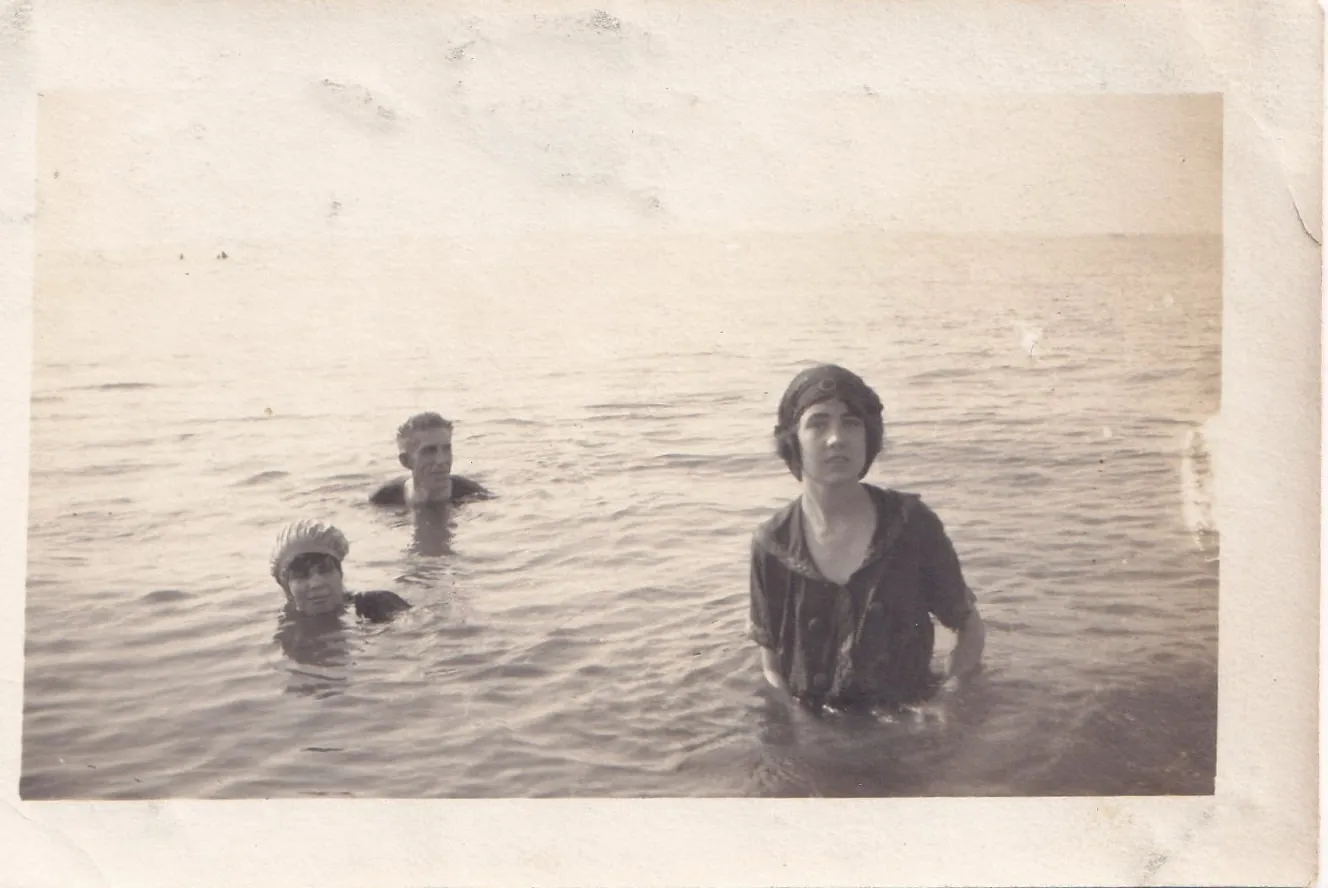 Antique black and white photograph of two women in swim caps and a man standing waist-deep in choppy open water under a grey sky