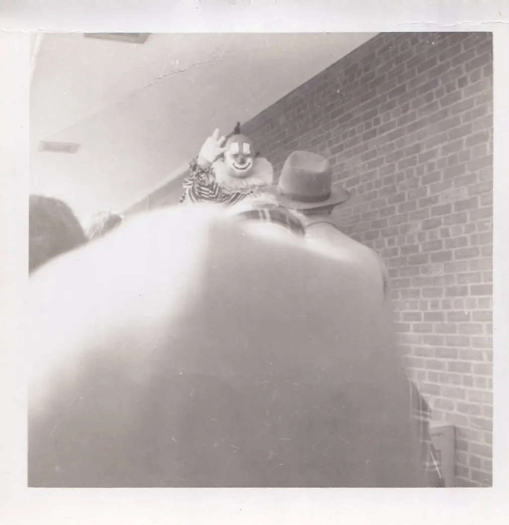 Black and white photograph of a costumed clown in a striped suit waving at the camera, nearly obscured by a large blurred figure in the foreground, a man in a fedora visible beside the clown against a brick wall