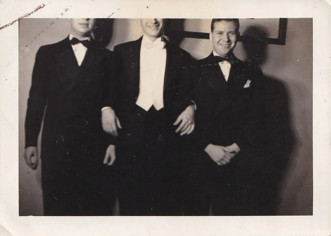 Vintage black and white photograph of three young men in formal evening wear — tuxedos and bow ties — posing together, slightly out of focus, a framed picture on the wall behind them