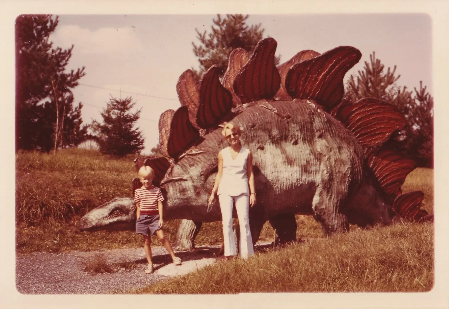 Found color snapshot of a woman and a young child standing beside a large life-size stegosaurus sculpture at an outdoor roadside attraction, pine trees visible behind, circa 1970s